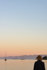 A gray haired woman watching the sailboats in the bay during sunset. She is seated on driftwood with her back to the camera, wearing a straw hat with a blanket over her shoulders.