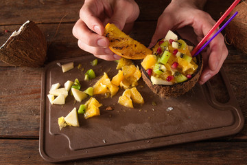 Decoration of tropical fruit salad in coconut shell. Apple, orange, kiwifruit and pomegranate ingredients are mixed in nut bowl with straws