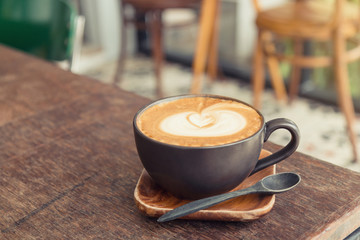 Hot latte coffee with latte art in a matt black cup on the wooden table in a coffee shop background (Vintage filter effect).