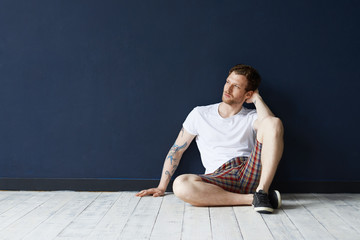 Fototapeta premium Picture of attractive young unshaven Caucasian man with tattoos wearing black sneakers, t-shirt and shorts having rest indoors, thinking about his life, future plans, looking thoughtful or dreamy