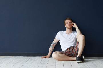 Indoor shot of handsome bearded young tattooed male of European appearance relaxing at home, sitting on floor wearing stylish clothes. People, lifestyle, rest, leisure and relaxation concept