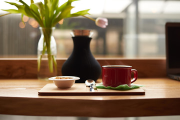 Mock up of hot coffee latte or cappuccino in red cup and white bowl with cane sugar cones resting on wooden table with decorative plants by the window glass in cozy restaurant during breakfast