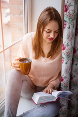 Young woman sitting at window drinking coffee and reading a book
