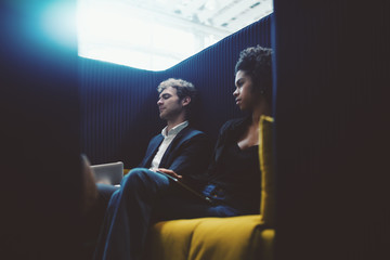 View through door aperture of black young businesswoman and caucasian man entrepreneur sitting both on greenish sofa in chill out recreational area of modern office and having rest