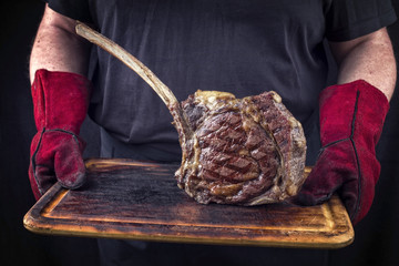 Man is holding Barbecue Wagyu Tomahawk Steak on old burnt cutting board in his hands with gloves