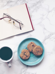 Black coffee and open notebook or journal with glasses and crunchy chocolate chip cookies or biscuits on a decorative plate on marble background. Flat lay. Top view. Copy space