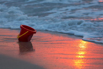 The sea shore at dawn, a baby bucket on the sand, the red sun reflects on the sand and in the water