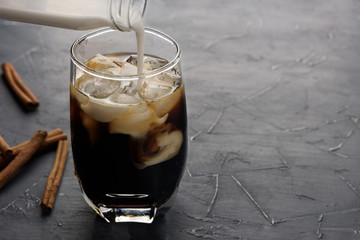 Cup of cold coffee on dark background. Iced espresso in a glass with cinnamon sticks on grey stone table. Pour cream in coffee. Copy space