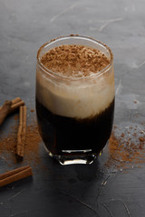 Cup of cold coffee on dark background. Iced espresso with cream in a glass with cinnamon sticks on grey stone table. Copy space