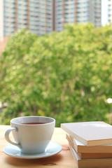Cup of hot tea with stack of books on wooden table by the window, with Blurred Background of green tree and high building