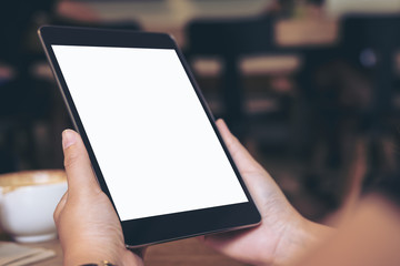 Mockup image of hands holding black tablet pc with blank white screen with coffee cup on wooden table in cafe