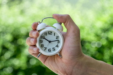 hand holding a white alarm clock  as business or time countdown concept with green bokeh in summer sun shine