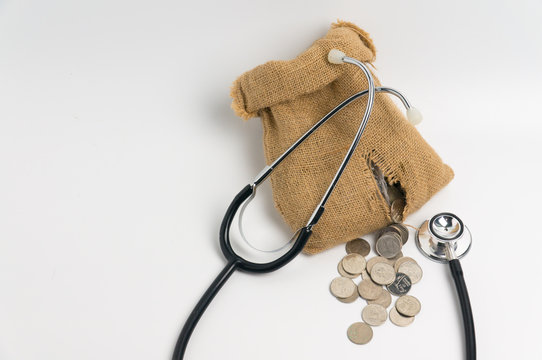 Damaged Money Bag With Coins Leaks From The Bag With Stethoscope Isolated On White Backgroudn, Concept Of Expenses Of Medical Treatment.