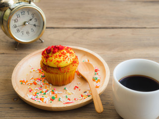 Yellow cupcakes put on a spherical wooden plate. Beside of cupcake have Vintage alarm clock and white coffee mug.All of it rests on wooden table.