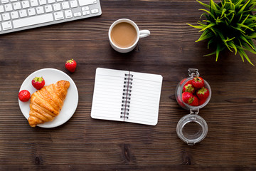 Light summer lunch at workplace. Coffee, strawberry, croissant near keyboard on wooden background top view copyspace