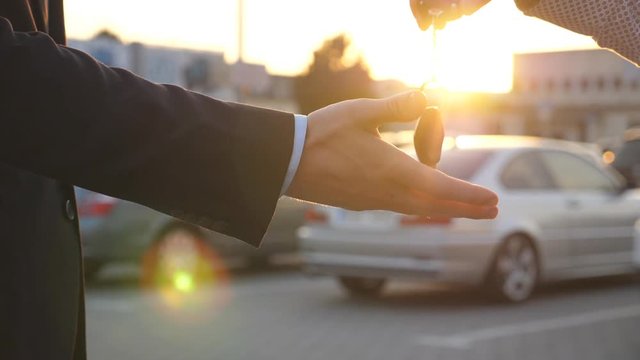 Arm of businessman passes car key at sunset. Male hands giving keys of car to his friend. Handshake between two business men outdoor. Close up Slow motion
