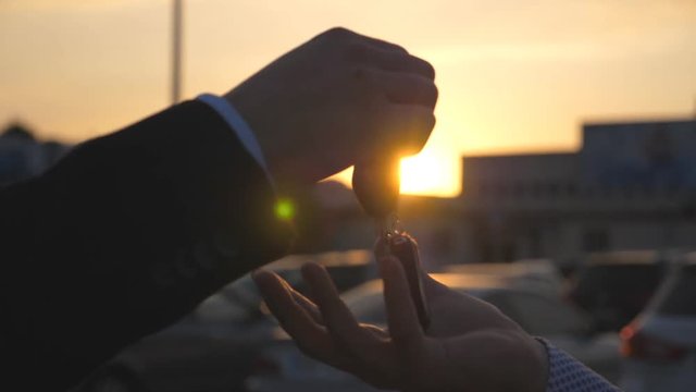 Male hands in suit giving keys of car to his friend at sunset time. Arm of businessman passes car key at evening. Handshake between two business men outdoor. Close up Slow motion