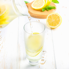 In a glass beaker, a cold lemonade is poured from a jug on a white wooden background surrounded by lemons.