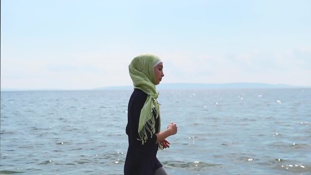 A young Muslim woman in a veil, which closes her hair, goes in for sports near water, the lady runs along the shore to improve endurance and strengthen the body