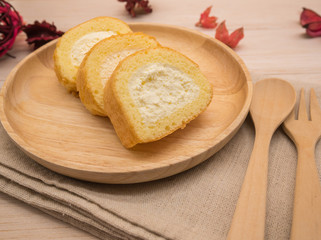 Naklejka premium Cake roll in wooden dish with wooden spoon and fork on beige wooden table,background and copy space decorated with red dried petals
