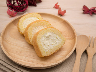 Cake roll in wooden dish with wooden spoon and fork on beige wooden table,background and copy space decorated with red dried petals