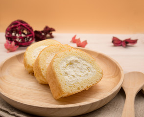 Cake roll in wooden dish with wooden spoon and fork on beige wooden table,orange background,copy space decorated with red flower petals