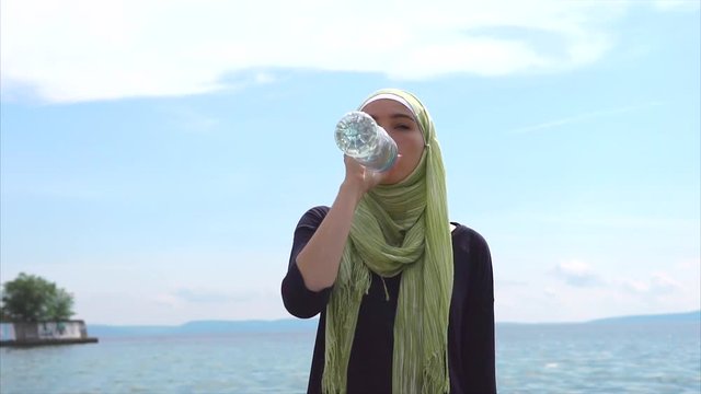 Modern Muslim in the headdress, which is called hijab drinks mineral water from a plastic bottle in nature, a lady in a scarf is near the river