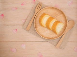 Top view cake roll in wooden plate with wooden fork,spoon  on brown fabric,beige wooden table,background with copy space,decorates with pink flower petals