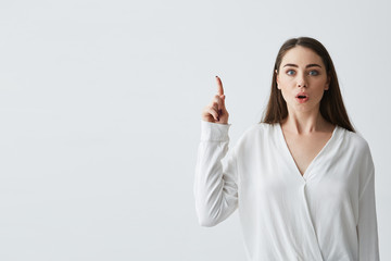 Fototapeta premium Surprised young beautiful businesswoman with opened mouth looking at camera pointing finger up over white background.