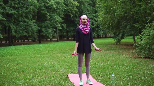 A young Muslim woman in closed clothes and a burqa doing exercises to strengthen her body in a summer park, a lady holds dumbbells and strengthens her arm muscles