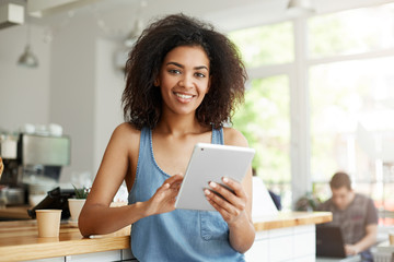 Young beautiful happy african girl resting in cafe smiling looking at camera holding tablet.