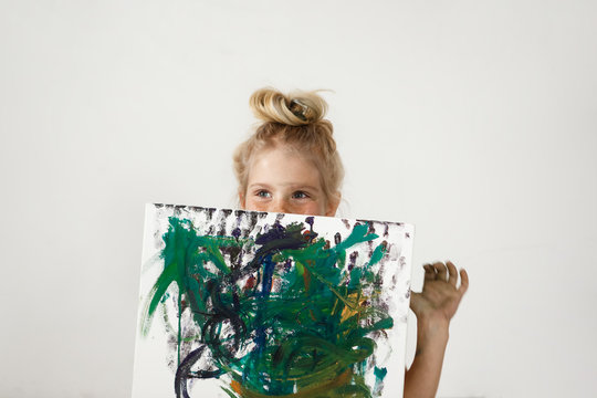Small European blonde girl with blue eyes and hair bun holding colourful picture and hiding her face. Happiness and joy of little girl is so charming. Kids` art activities.