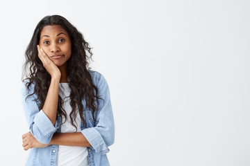 Human emotions, feelings, reaction and attitude. Attractive dark-skinned girl in denim shirt with long wavy hair keeping hand on cheek in doubt and suspicion, feeling sceptical about something.
