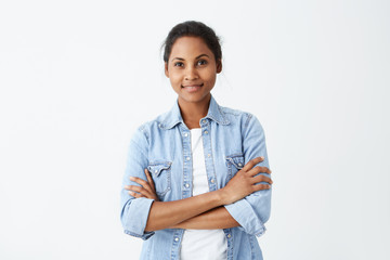 Photo of restful African-american woman with black hair standing crossed hands having sincere and delightful smile posing on white studio wall. Happy dark-skinned female rejoicing her life.
