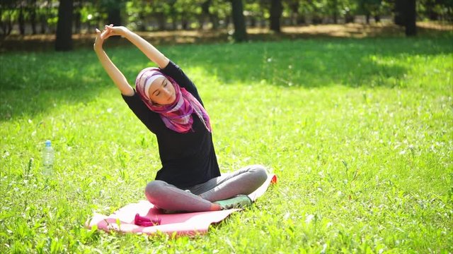 A young Muslim woman in a scarf, called a burqa, does stretching the back muscles, the lady is in the summer park and is doing sports