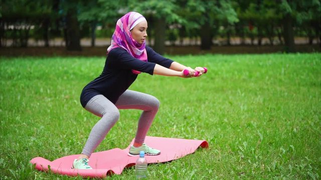 A modern Muslim woman who clothed a veil over her head is engaged in sports on a summer day in the park, the lady does exercises to strengthen the leg muscles and stretch the back of the thigh