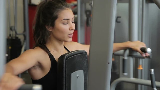 Woman athlete doing lateral raise machine in gym indoors. holding elbows at shoulder level and returning to starting position. Attractive brunette dressed in black compression top with neckline is