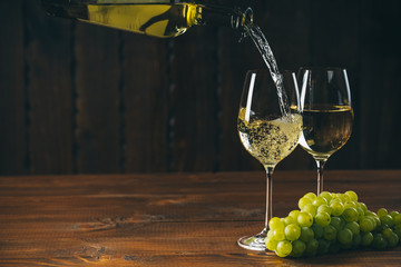 Pouring white wine into a glass with a bunch of green grapes against wooden background