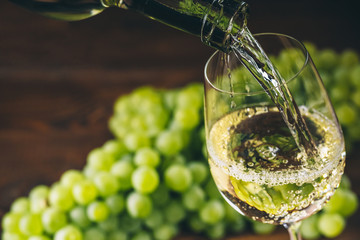 Pouring white wine into a glass with a bunch of green grapes against wooden background - obrazy, fototapety, plakaty