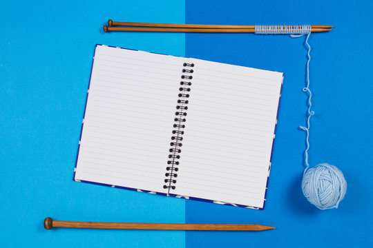 Knitting needles, blue yarn ball and open paper notebook on blue background