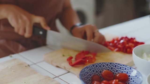 Hand held shot of young woman chef chopping fresh organic paprika peppers full of red color, on beautiful designer kitchen counter and artisan wooden cutting board