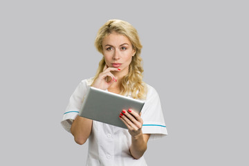 Portrait of young nurse with digital tablet. Thoughtful young female doctor or nurse holding computer tablet and having an idea, grey background.