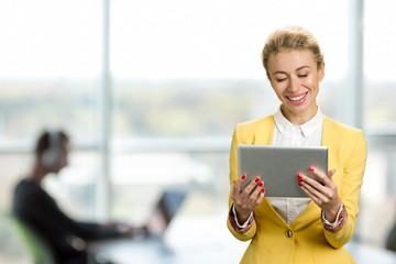 Smiling business lady with pc tablet. Cheerful young business woman looking on computer tablet and smiling, office window background.