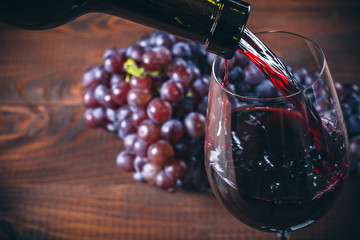 Pouring red wine into the glass with a bunch of red grapes against wooden background