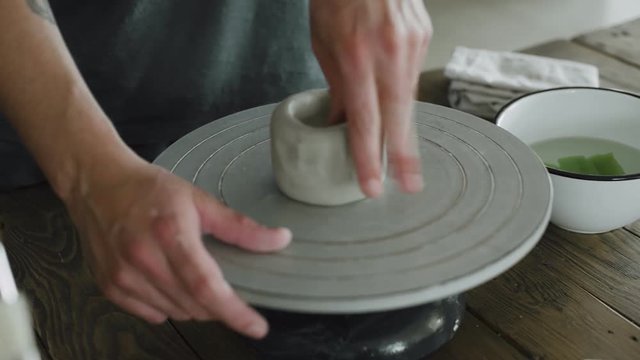 Close up view of man's hands making ceramic pot of raw grey clay on pottery wheel in his home studio.
