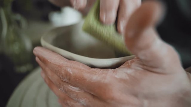 Close up view of artist man making ceramic pot of raw grey clay on pottery wheel and smoothing edges with wet sponge in workshop.