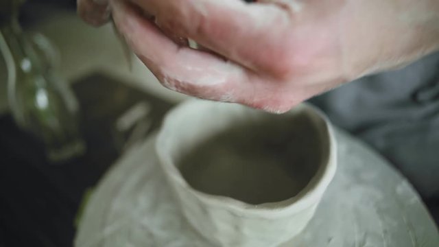 Ceramist man works on pot of raw grey clay and decorate it with little detail nose to make stylisation of face in his pottery studio. Close up view.