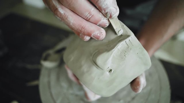 Artist man decorates freshly hand made raw clay pot with nose detail for stylisation of face using wooden stick tool in workshop.