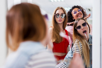 Trendy female friends trying on stylish sunglasses looking in mirror, smiling, having fun in accessory store