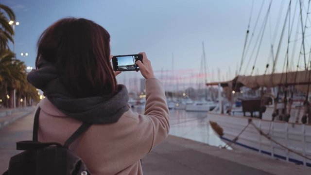 Back view on brunette girl is taking photos of turistic view of marina with many parked sail boats at early morning before sunrise.Peaceful romantic light with pinky clouds and fresh breeze from sea.